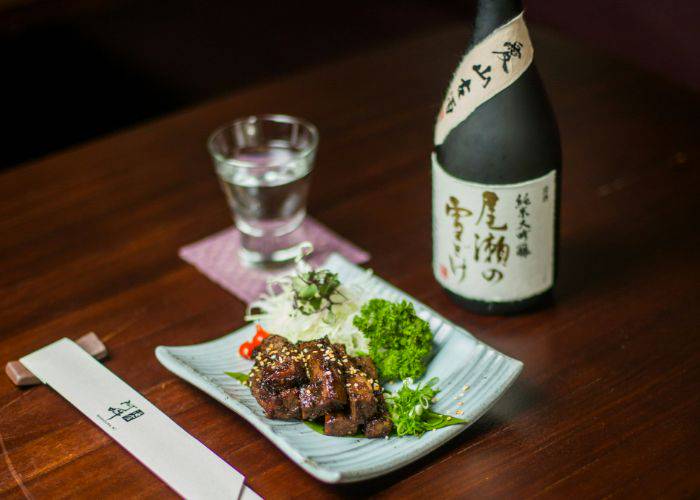 A black bottle of sake, a poured cup of sake and a plate of saucy meat.